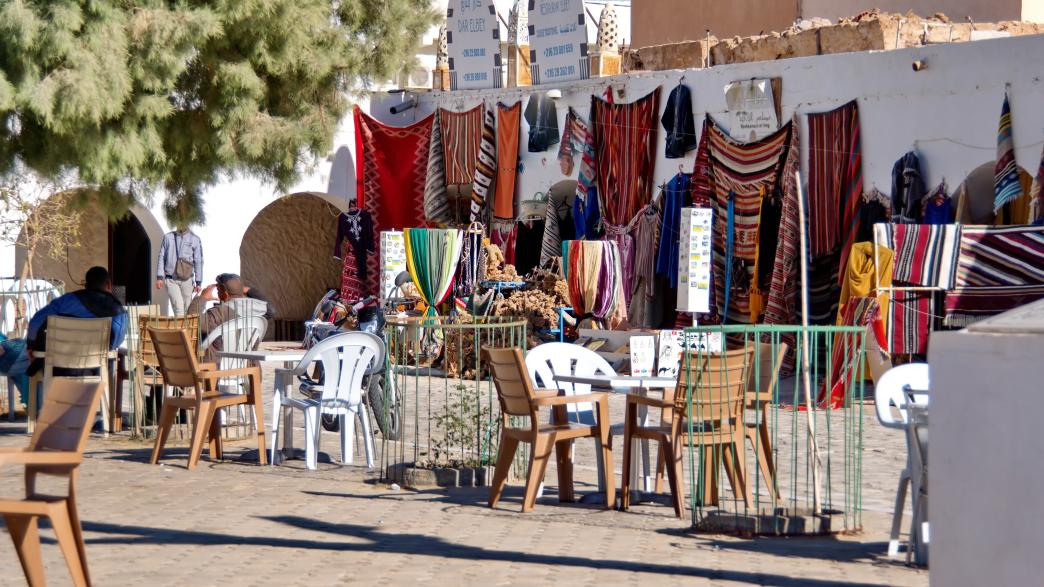 Cafe, with tables and chairs in the courtyard, set up in front of a stall selling colorful textiles in the market