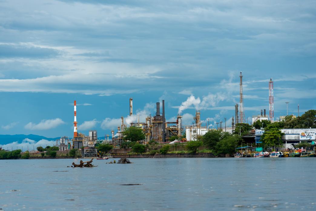 Industrial oil refinery complex with chimneys emitting smoke, located on the banks of a river with boats docked in the foreground, under a cloudy blue sky.